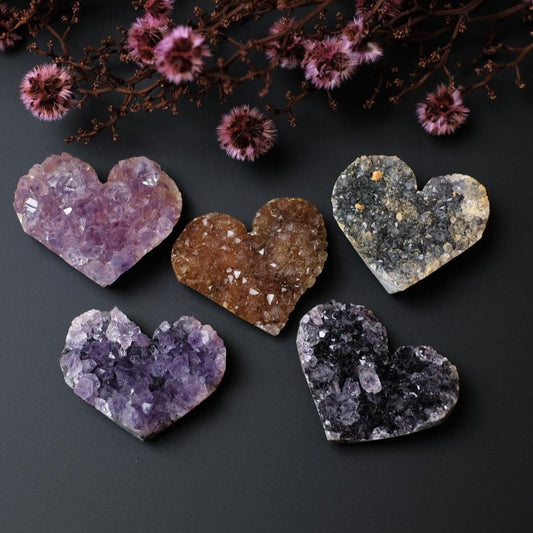 A collection of heart-shaped druzy Amethyst crystals displayed on a dark surface with some dried flowers in the background.