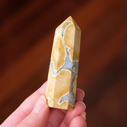 A hand holding a polished natural crystal point with a yellow and gray agate-like pattern, set against a blurred dark brown wooden background.