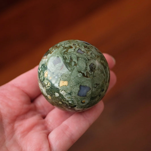 A hand holding a polished green stone sphere with natural inclusions and flecks, set against a blurred warm brown background.